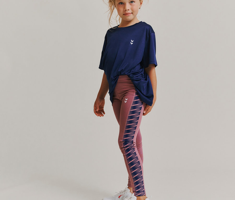 Mädchen in dunkelblauem T-Shirt und rosa Leggings mit blauen Streifen steht vor weißem Hintergrund.