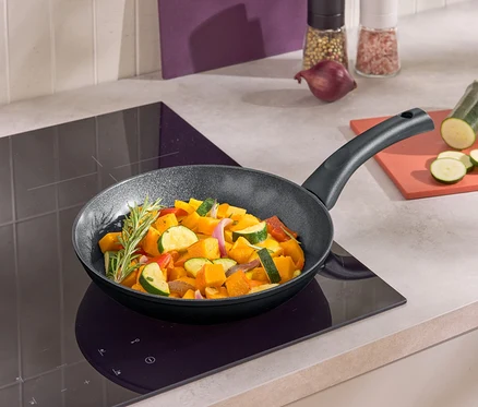 Eine Bratpfanne mit gebratenem Gemüse auf einem Induktionskochfeld. Neben der Pfanne liegt eine geschnittene Zucchini auf einem Brettchen.