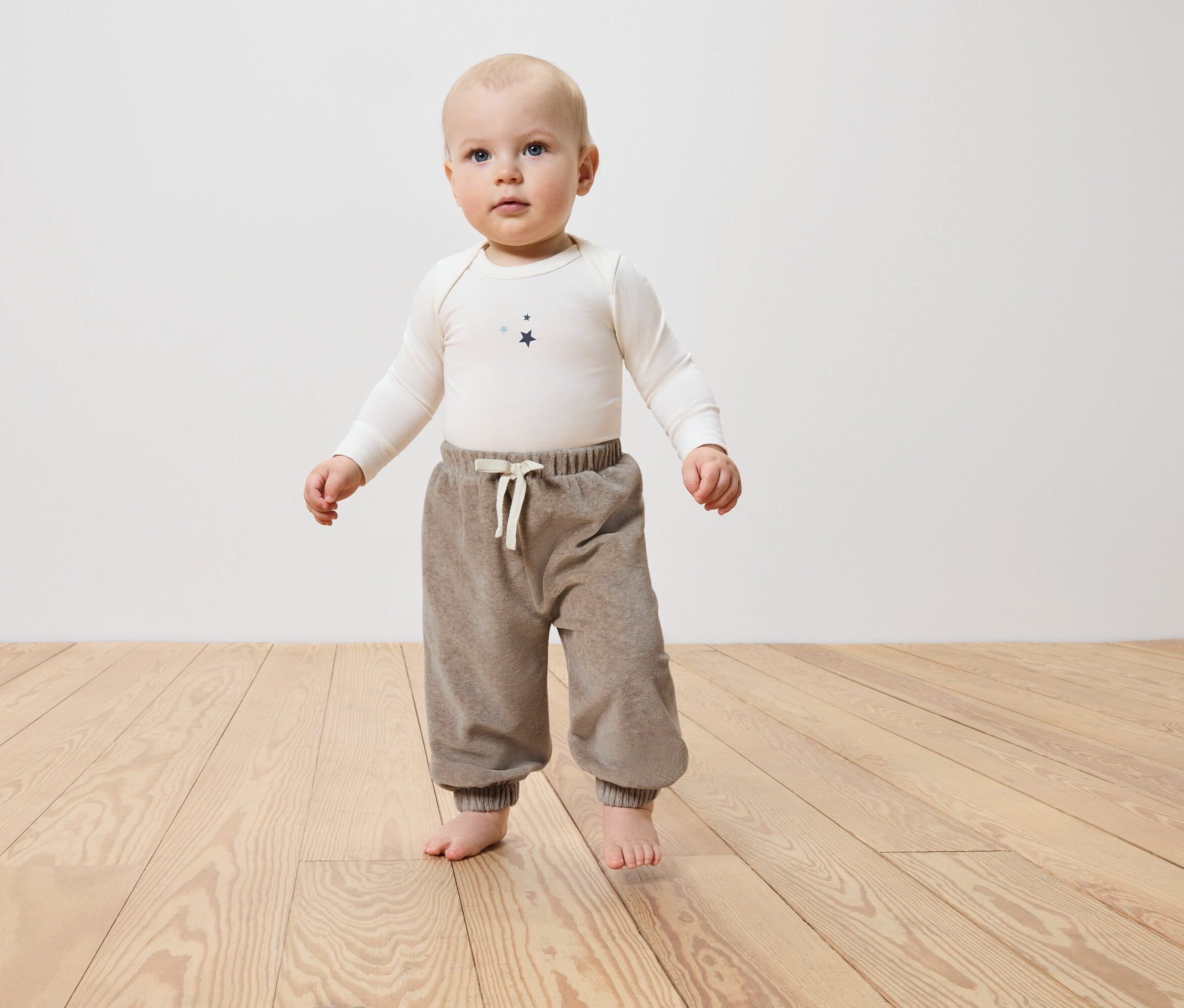 Ein Baby steht barfuß auf einem Holzboden und trägt eine braune Hose und ein weißes Langarmshirt.