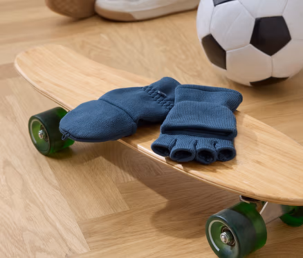 Blaue Fingerhandschuhe liegen auf einem hölzernen Skateboard mit grünen Rädern. Im Hintergrund befinden sich ein Fußball und Schuhe.