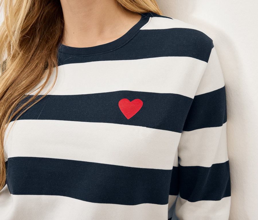 Detailaufnahme vom Oberkörper einer Frau, die ein gestreiftes, langärmeliges Shirt mit einem kleinen roten Herz auf der Brust trägt.