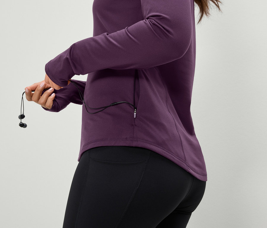 Frau hält Kopfhörer, trägt eine violette Sportjacke und schwarze Leggings.