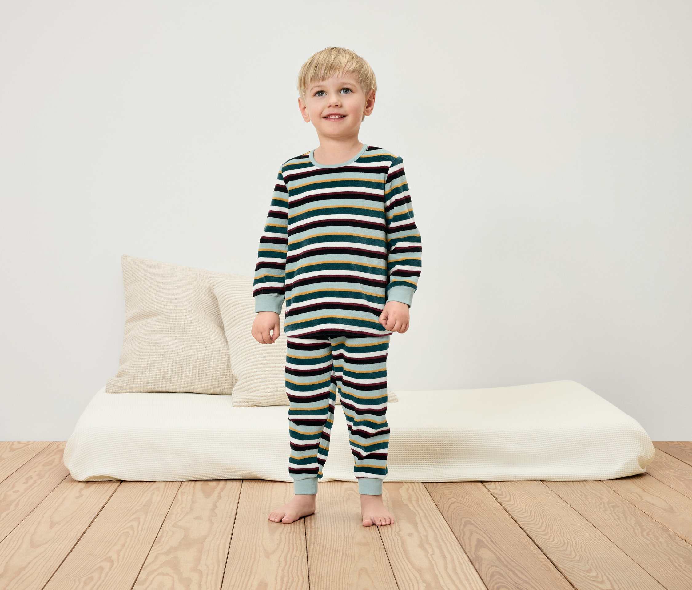 Ein Junge im gestreiften Pyjama steht barfuß auf einem Holzboden vor einer weißen Wand.