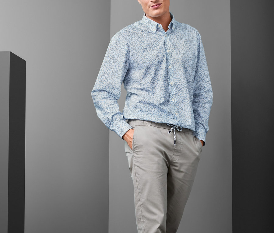 Mann posiert und trägt ein hellblaues Hemd mit Button-down-Kragen in Comfort Fit, graue Hosen und blaue Turnschuhe.