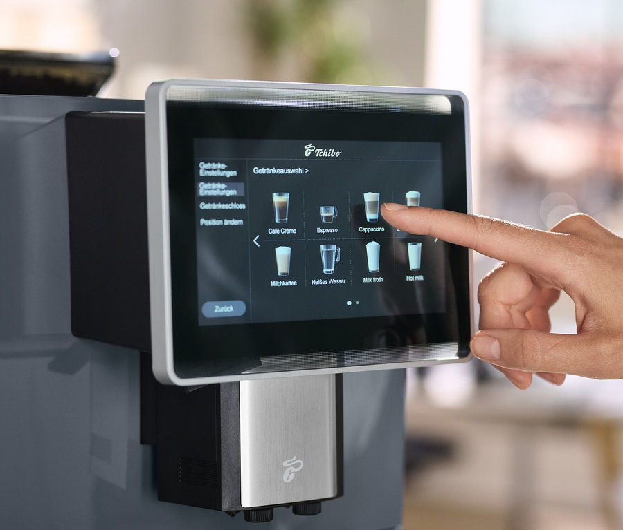 Ein Finger berührt den Bildschirm einer Tchibo Kaffeemaschine, auf dem eine Getränkeauswahl angezeigt wird.