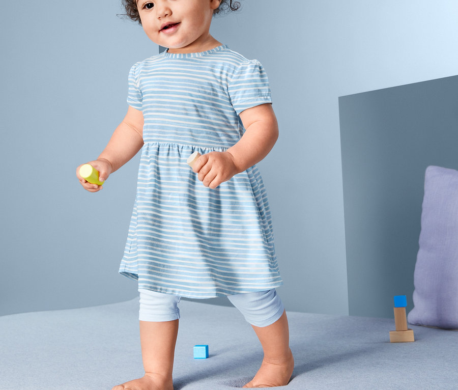 Ein Mädchen in einem blau-weiß gestreiften Baumwollkleid (2 Stück) steht auf einem Bett.