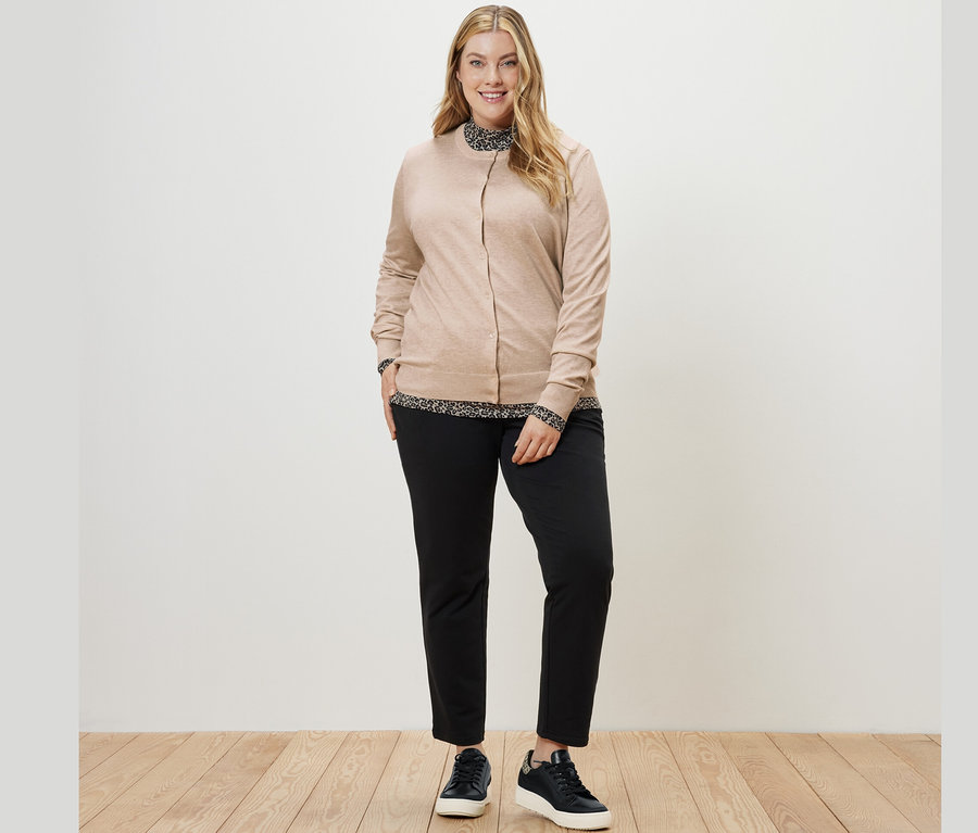 Eine Frau trägt ein bedrucktes Blusenshirt mit Stehkragen, einen Feinstrick-Cardigan und eine schwarze Joggpants.