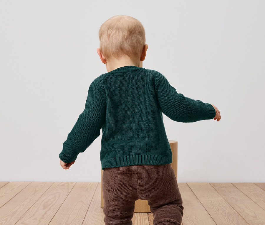 Kleinkind in grünem Pullover und braunen Leggings steht mit dem Rücken zur Kamera.