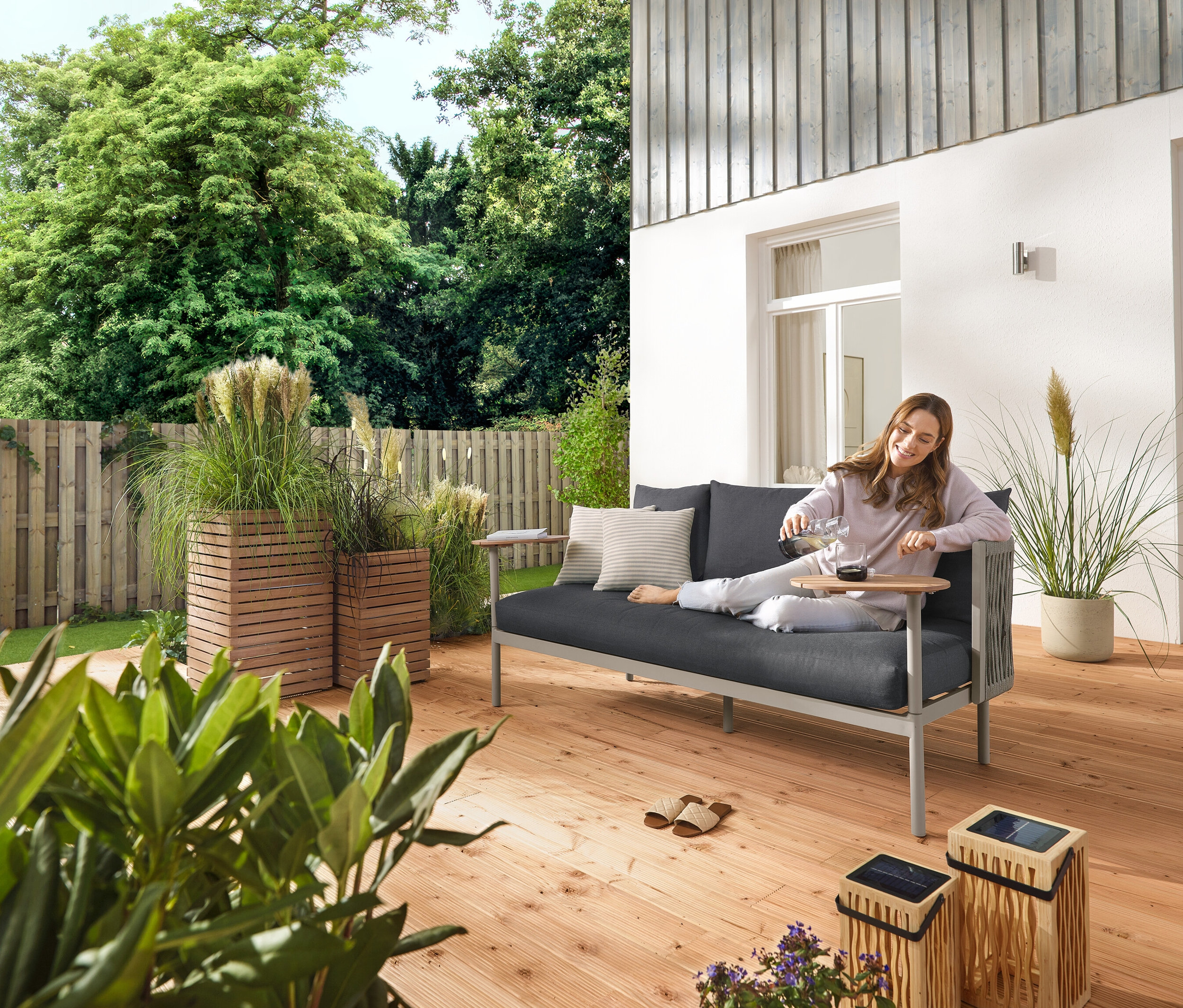 Eine Frau gießt draußen auf einer Gartenterrasse Wasser in ein Glas, neben ihr stehen eine große und eine kleine Bambus-Solar-Laterne, sowie 5 LED-Solar-Spieße und eine Gartensofa mit 2 Drehtischen. Außerdem sind 2 Pflanzsäulen »Tinus« zu sehen.