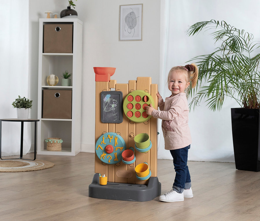 Mädchen spielt mit Smoby Life Activity Wall, einem Spielzeug mit Tafel, Drehscheibe und Behältern. Regal und Pflanze im Hintergrund.