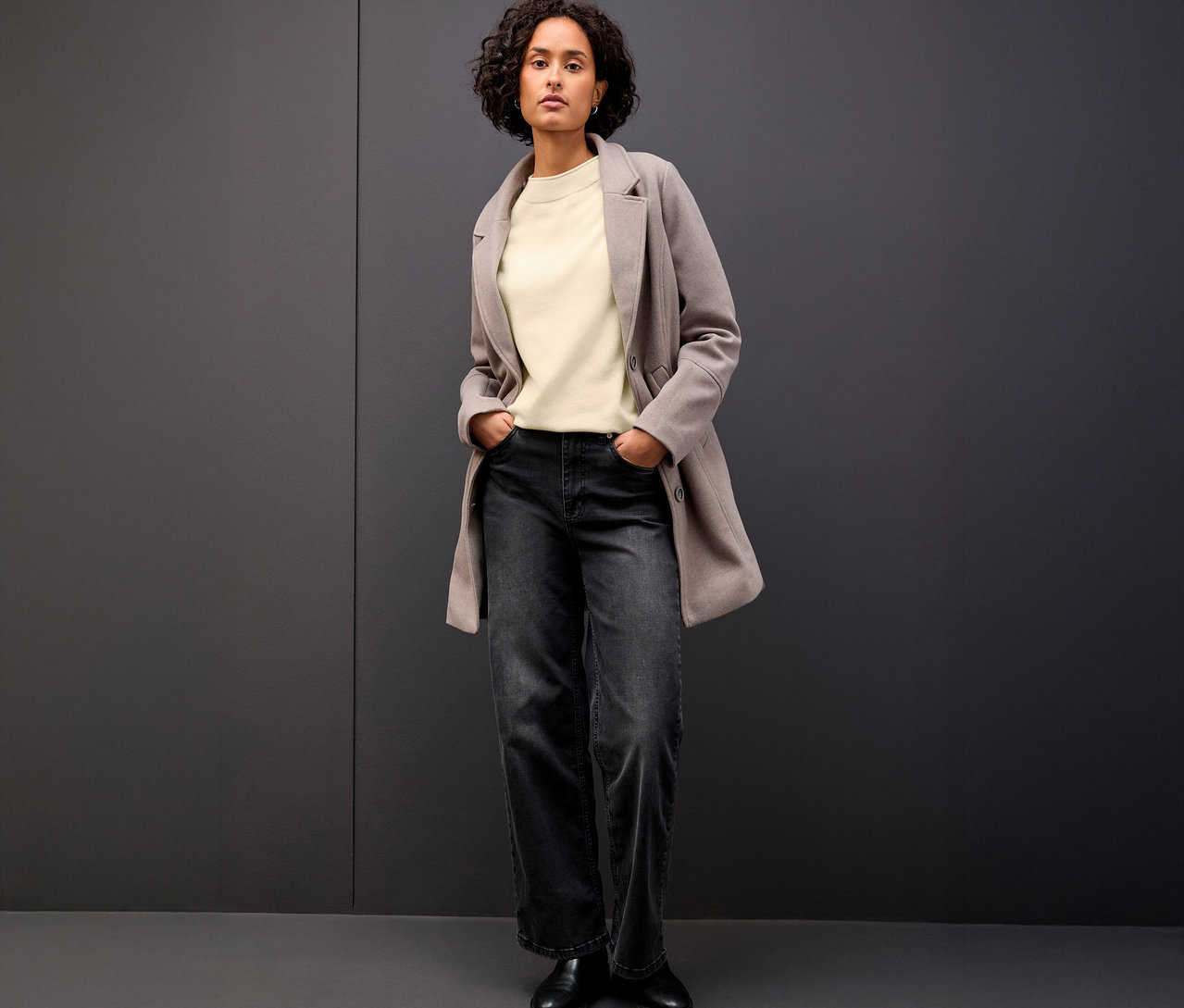 Frau steht mit den Händen in den Hosentaschen und trägt einen beigen Street One Strickpullover mit Rollkante, eine graue Jacke und eine dunkle Jeans.