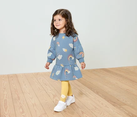 Ein kleines Mädchen mit lockigem Haar trägt ein blaues Kleid mit Regenbogenmuster, gelbe Leggings und weiße Schuhe.