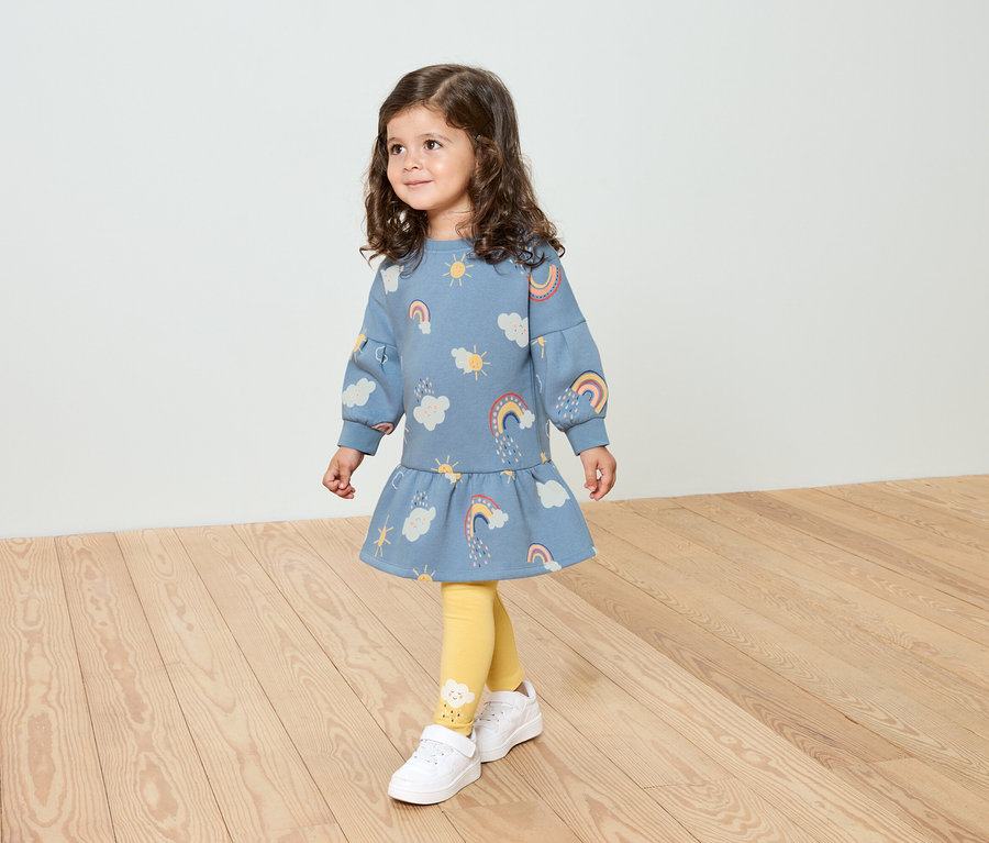 Ein kleines Mädchen mit lockigem Haar trägt ein blaues Kleid mit Regenbogenmuster, gelbe Leggings und weiße Schuhe.