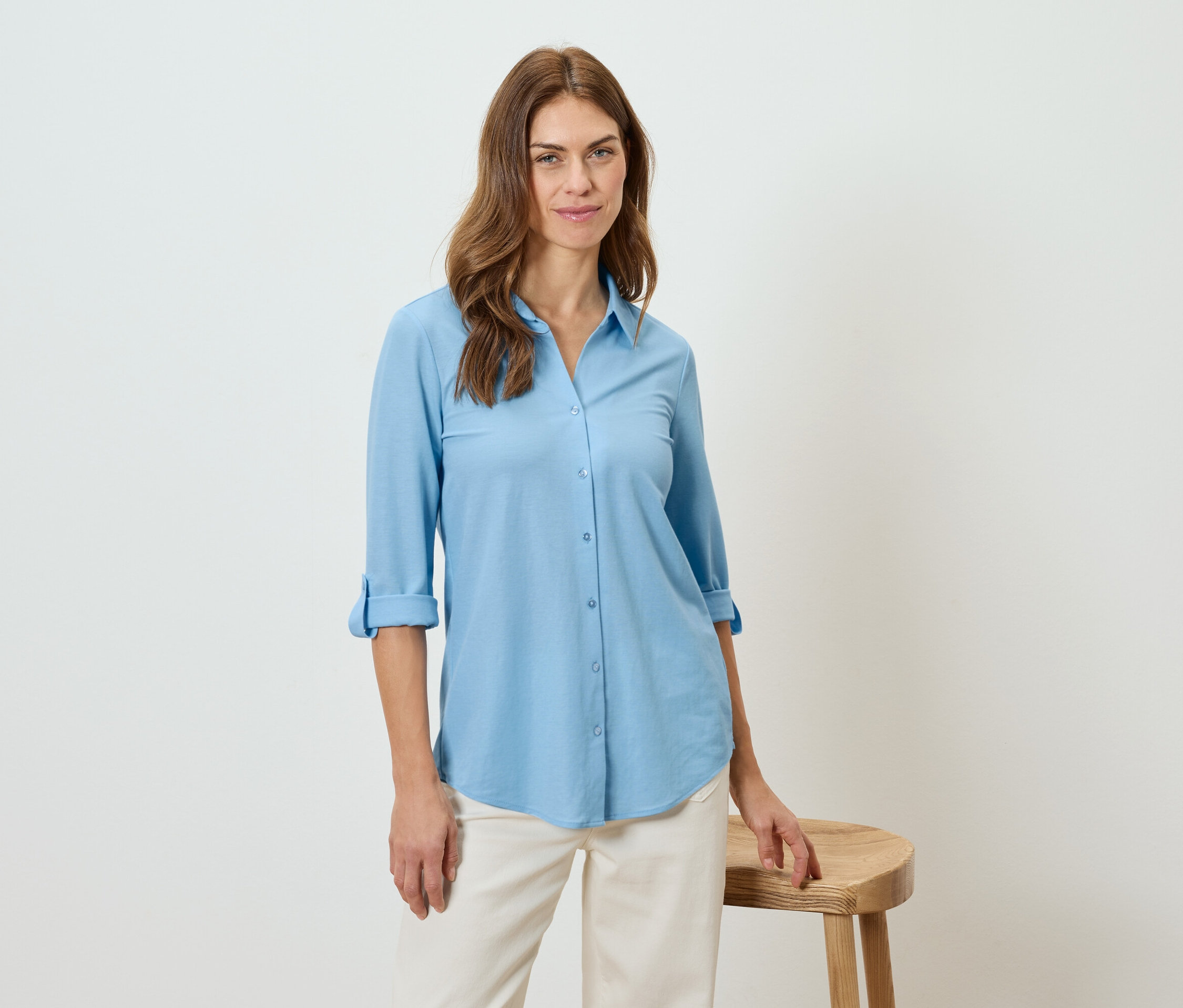 Frau trägt ein hellblaues Hemd und beigefarbene Hosen, lehnt an einem Hocker.