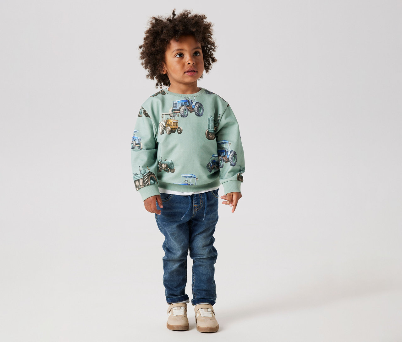 Junge mit Afro trägt einen mintfarbenen Pullover mit Traktoren und NAME IT Kinder-Sweat-Jeans und beigefarbene Schuhe.