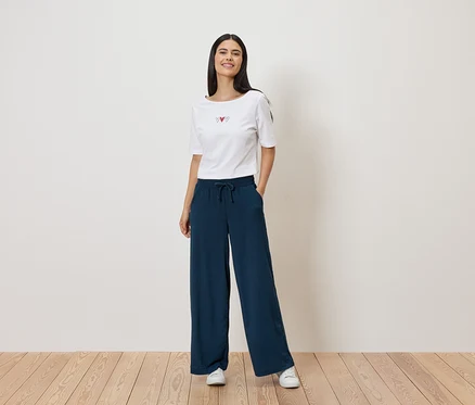 Frau mit weißem T-Shirt mit Herz-Motiv und weiter, dunkelblauer Hose mit Kordelzug. Sie trägt weiße Turnschuhe.
