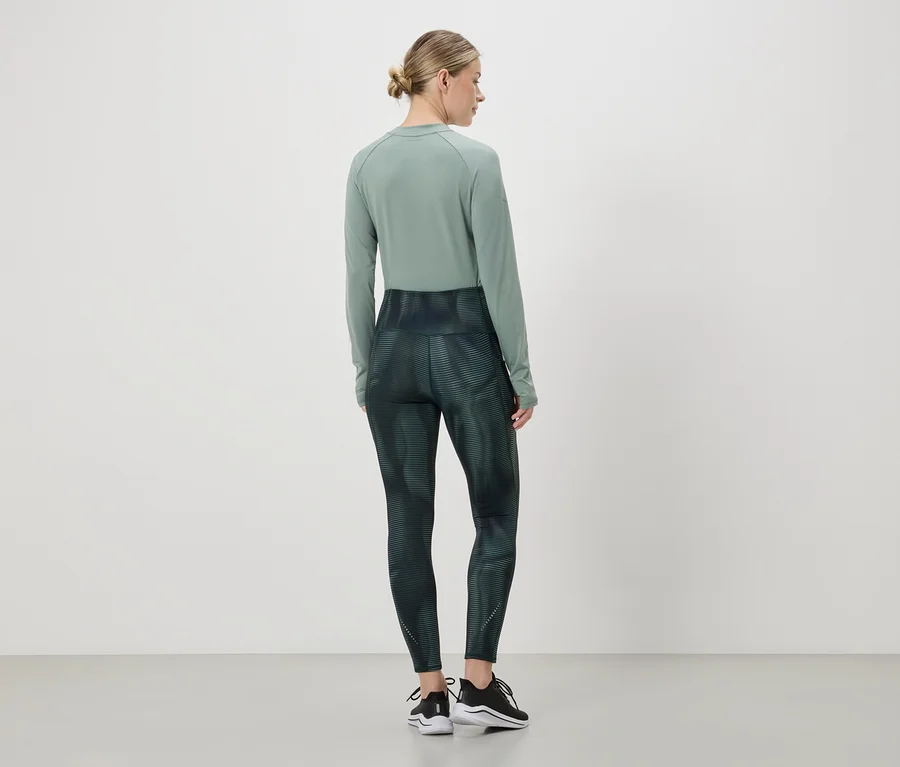 Eine Frau mit Dutt trägt ein grünes Langarmshirt und Leggings mit abstraktem Muster, dazu schwarze Sneaker.