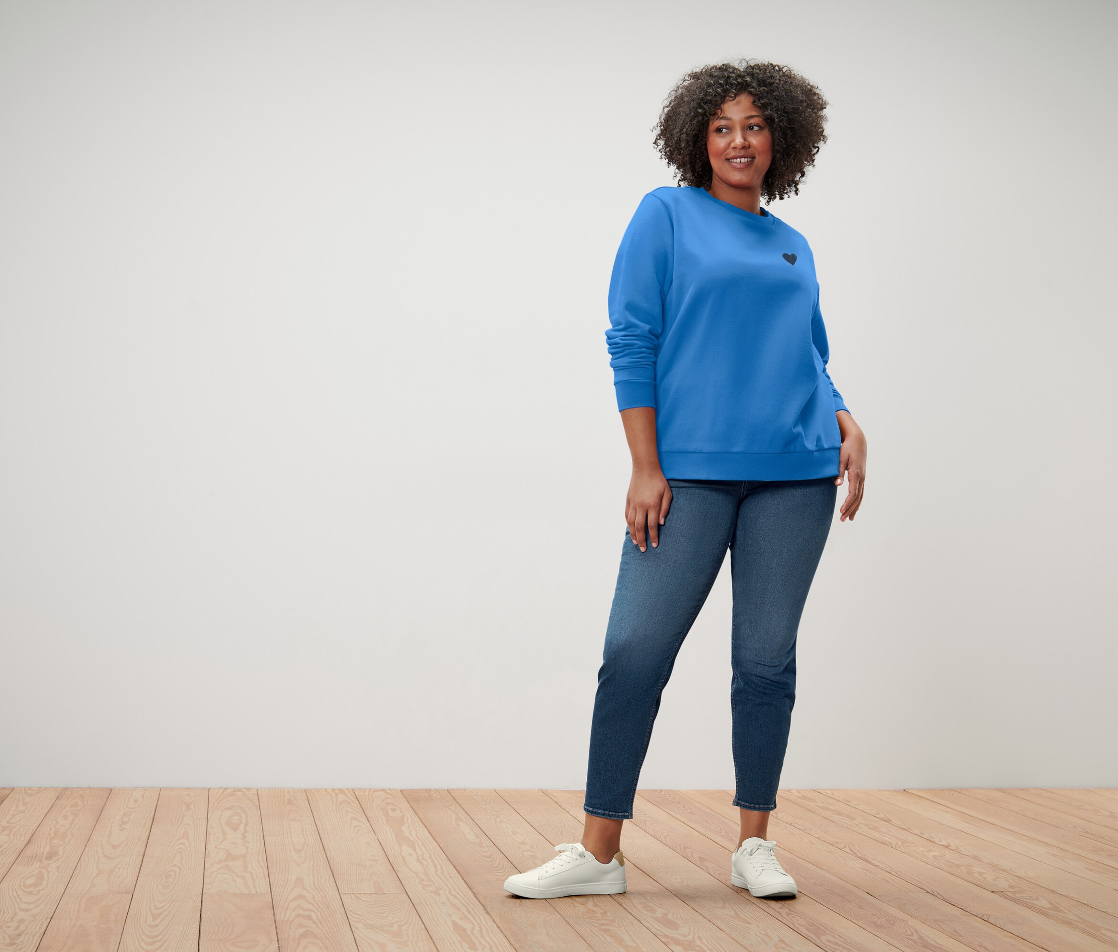 Frau mit lockigem Haar trägt einen blauen Pullover mit einem kleinen schwarzen Herz, blaue Jeans und weiße Turnschuhe.