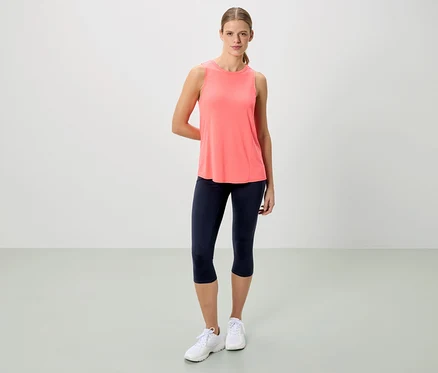 Frau steht in korallfarbenem Tanktop, marineblauen Capri-Leggings und weißen Turnschuhen vor einem neutralen Hintergrund.