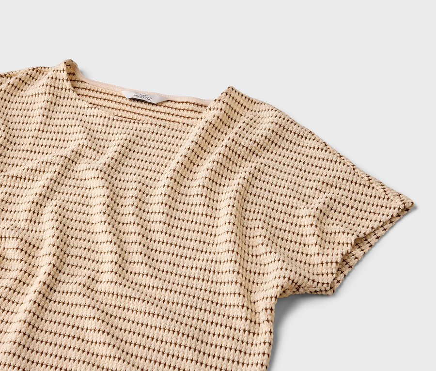 Nahaufnahme eines kurzärmeligen Oberteils in Beige mit braunem Punktmuster auf grauem Hintergrund.