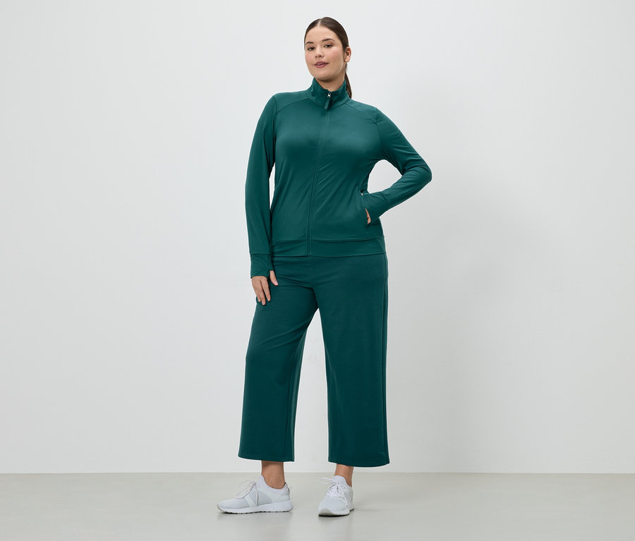 Frau in smaragdgrünem Trainingsanzug mit Reißverschlussjacke und Culottes, dazu weiße Turnschuhe.