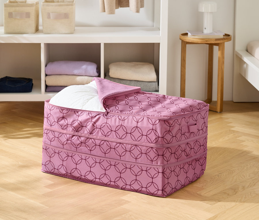 Auf einem Holzboden steht eine rosafarbene Aufbewahrungstasche mit geometrischem Muster, teilweise mit einer weißen Decke gefüllt.