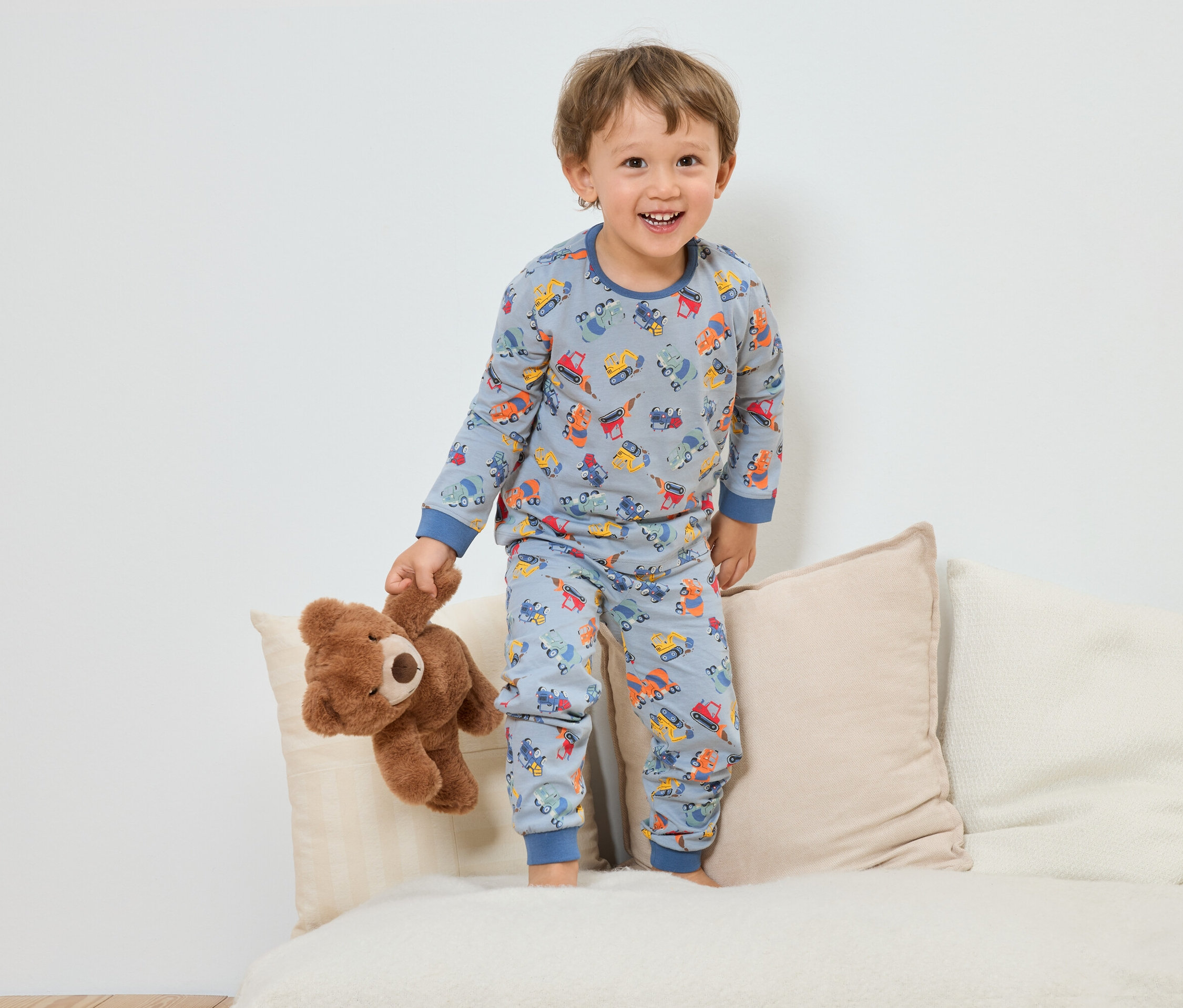 Ein lächelnder Junge im blauen Schlafanzug mit Baufahrzeugmuster steht mit einem Teddybär auf einem Sofa.