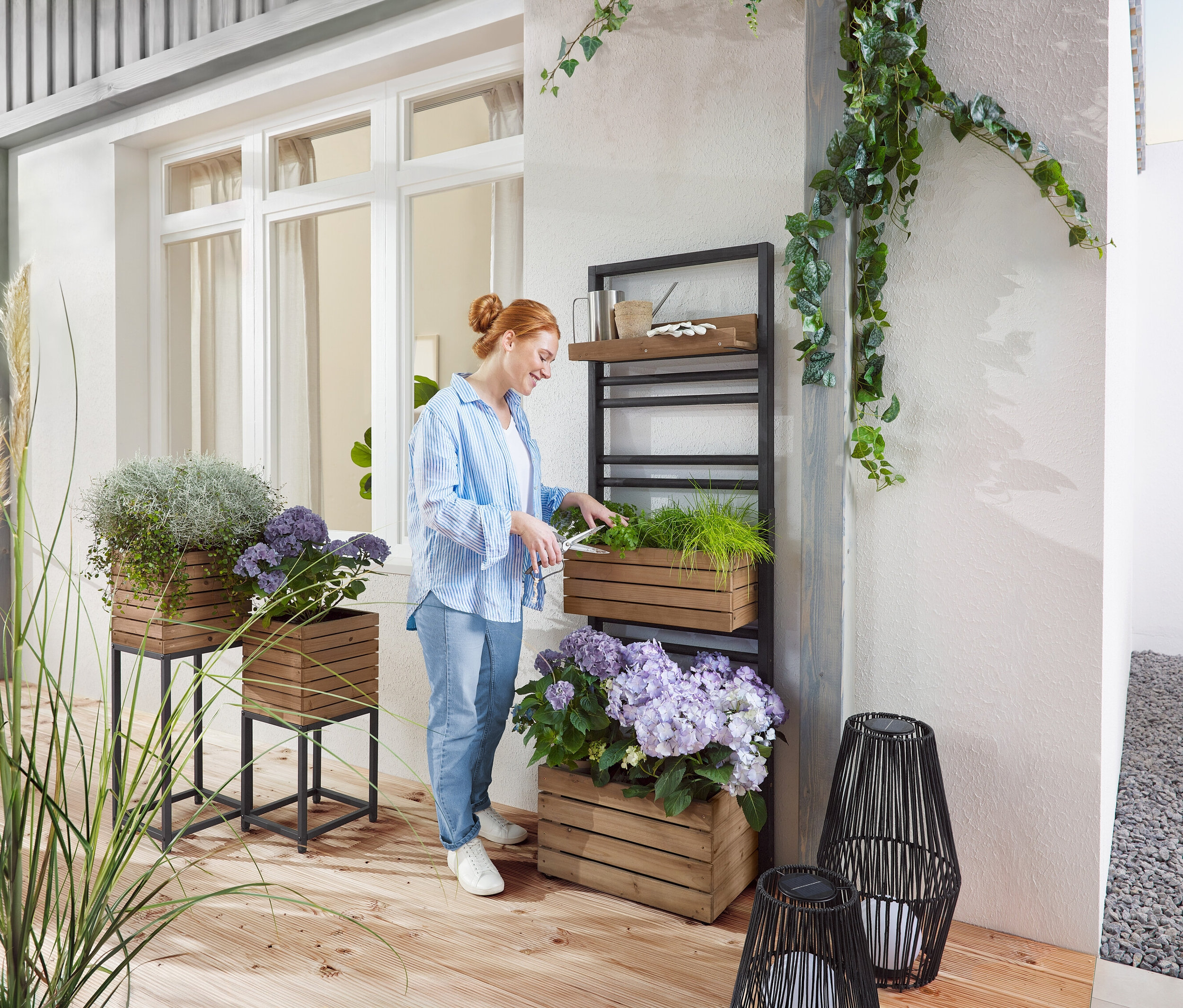Frau schneidet Grünpflanze in Holzkasten auf einem Regal. Draußen stehen Holzpflanzgefäße mit Pflanzen und lila Blumen.
