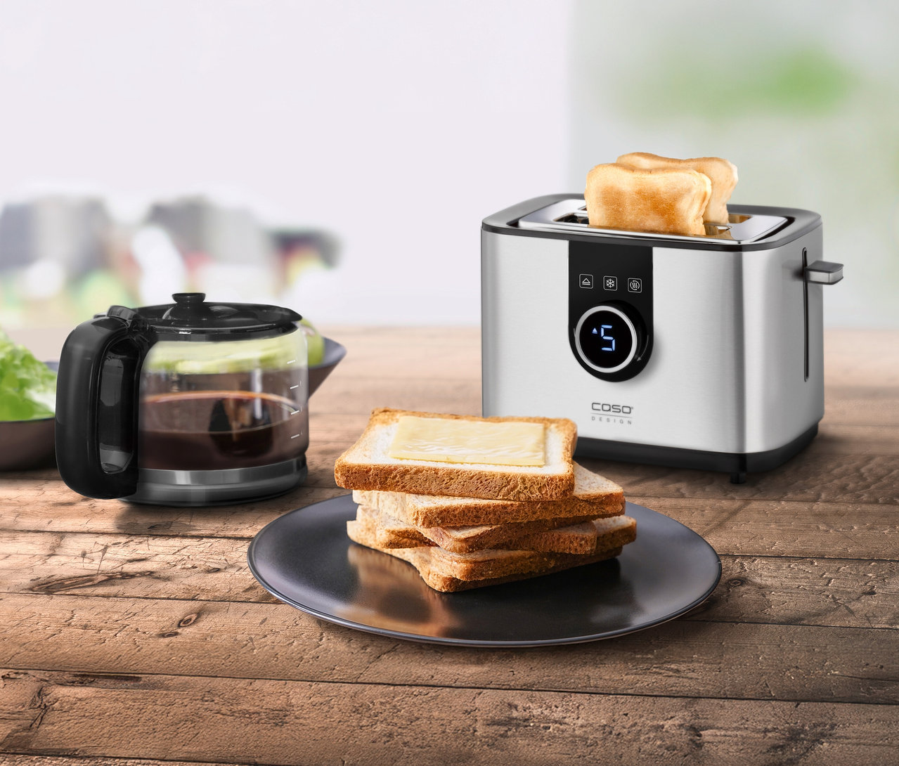 Ein silberner Toaster Selection T2 steht neben einem Stapel Toastbrot und einer Kaffeekanne auf einem Holztisch.