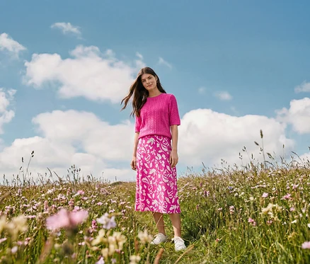 Eine Frau steht auf einer Wiese und trägt einen bedruckten Midi-Rock, Creolen Wave und Leder-Sneaker.