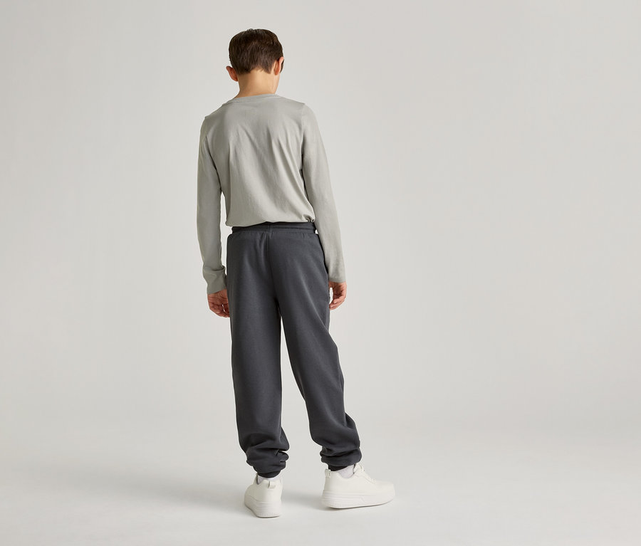 Ein Kind mit hellgrauem Langarmshirt und dunkelgrauer Jogginghose steht mit dem Rücken zum Betrachter vor einem hellen Hintergrund.