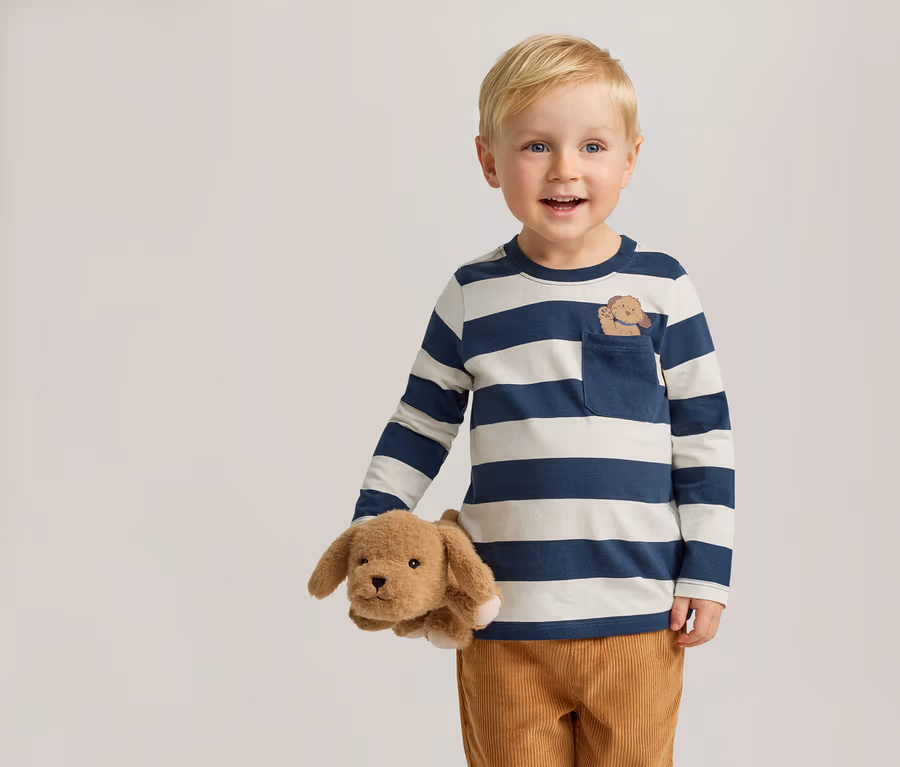 Lächelnder Junge mit blonden Haaren hält einen Stoffhund. Er trägt ein blau-weiß gestreiftes Langarmshirt mit einer Hundestickerei auf der Tasche.