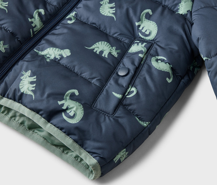 Nahaufnahme einer marineblauen, gesteppten Jacke mit hellgrünem Dinosaurier-Muster, Reißverschluss und einer Tasche mit Druckknopf.