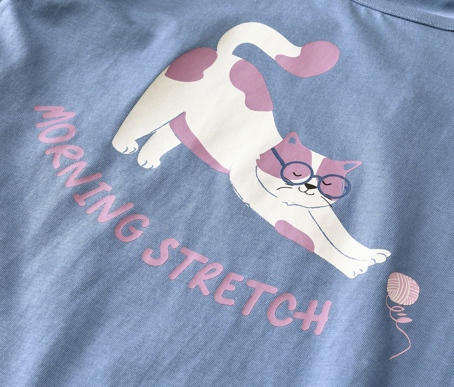 Hellblauer Stoff mit einem Aufdruck einer weißen Katze mit rosa Flecken und blauer Brille, die sich streckt. Rosa Text lautet "MORNING STRETCH" neben einem kleinen rosa Wollknäuel.