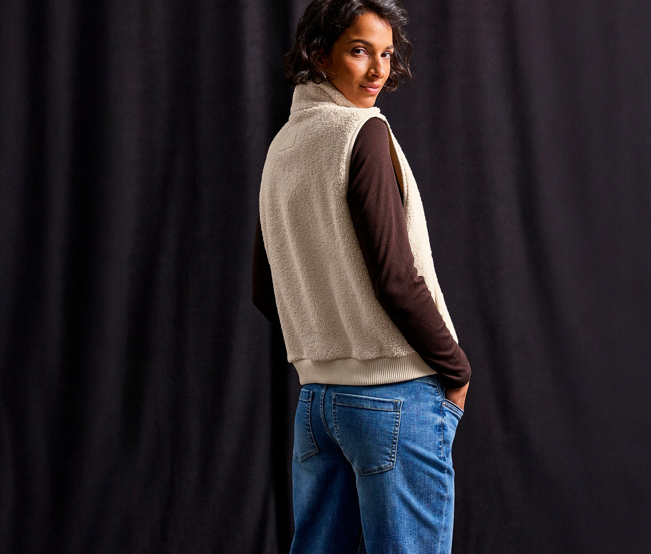 Frau mit braunem Oberteil und blauen Jeans, die eine Street One Teddyweste trägt und über die Schulter blickt.
