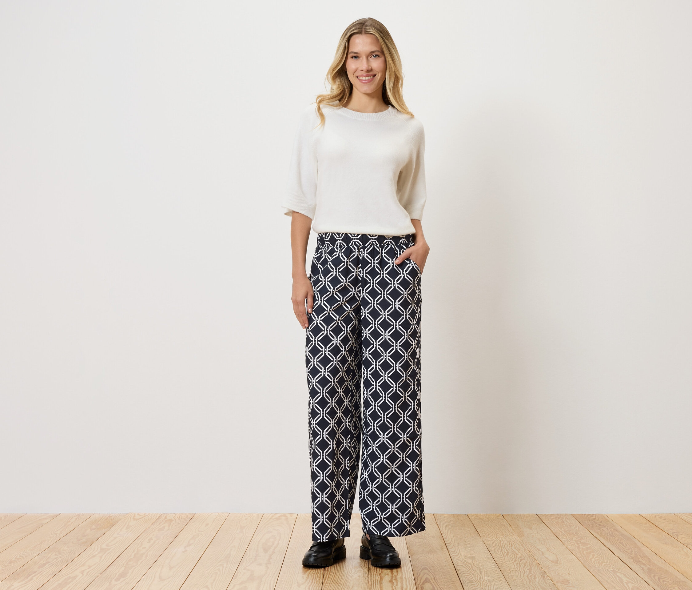 Frau mit weißem Oberteil und gemusterter Hose, die vor einer Wand auf Holzboden steht.