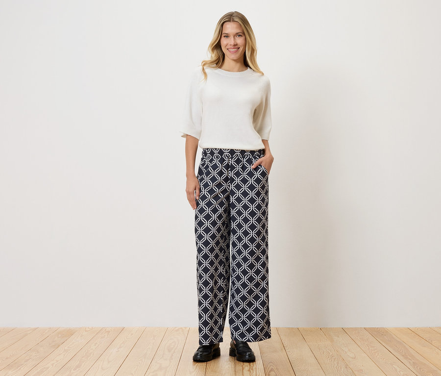 Frau mit weißem Oberteil und gemusterter Hose, die vor einer Wand auf Holzboden steht.