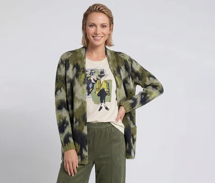 Lächelnde Frau mit kurzen blonden Haaren trägt eine grün gemusterte Strickjacke, ein helles T-Shirt mit Grafikdruck und olivgrüne Hosen.