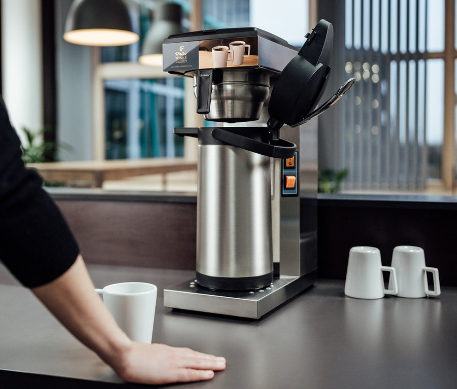 Eine Frau steht an einem Tisch mit einer Tchibo Businessline Profi-Filterkaffeemaschine ohne Kanne und einer Tasse.