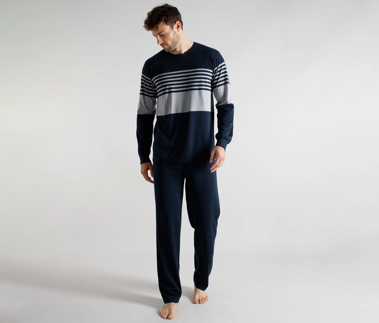 Mann trägt ein dunkelblaues und grau gestreiftes langärmeliges Pyjama-Oberteil und eine einfarbige dunkelblaue Pyjamahose, blickt nach unten.