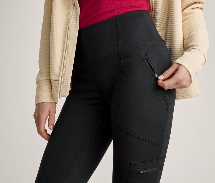 Eine Person in schwarzen Hosen mit Reißverschlusstaschen und einer beigen gerippten Jacke öffnet einen Reißverschluss an einer Seitentasche der Hose.