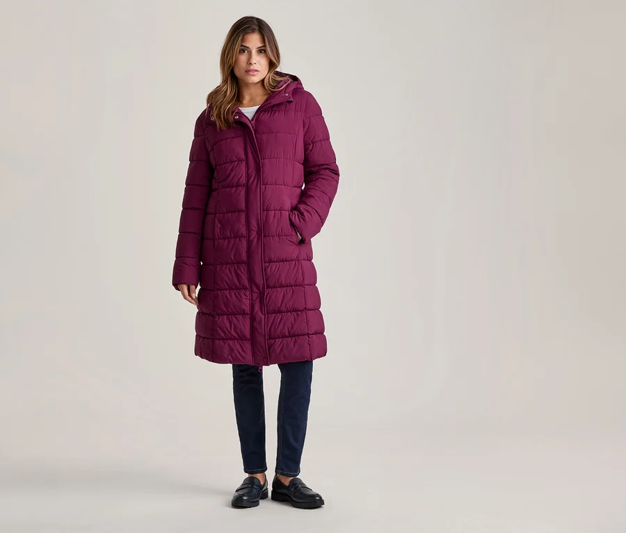 Frau steht in einem langen, lila Steppmantel mit Kapuze, dunklen Jeans und schwarzen Slippern, die Hände in den Taschen.