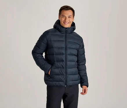 Lächelnder Mann in einer dunkelblauen Kapuzen-Steppjacke und schwarzen Hosen, eine Hand in der Jackentasche.