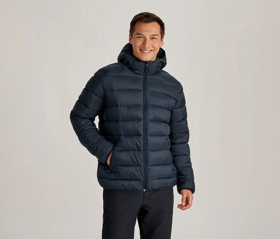 Lächelnder Mann in einer dunkelblauen Kapuzen-Steppjacke und schwarzen Hosen, eine Hand in der Jackentasche.