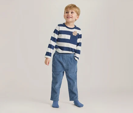 Junger Junge steht, blickt lächelnd nach oben und trägt ein blau-weiß gestreiftes Langarmshirt mit Tier-Brusttasche, blaue Cordhose und blaue Socken.