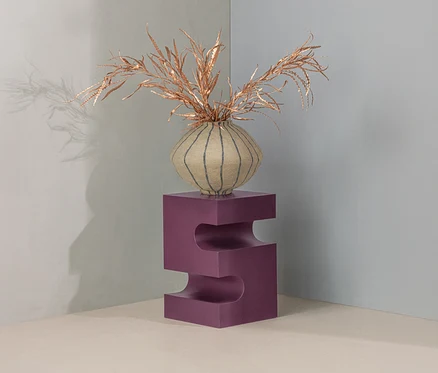 Eine runde, gestreifte beigefarbene Vase mit goldenen Trockenpflanzen steht auf einem lila S-förmigen Sockel in einer hellen Raumecke.