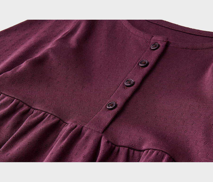 Nahaufnahme eines burgunderfarbenen Strickkleidungsstücks mit dezentem Texturmuster. Es hat einen Rundhalsausschnitt, eine Knopfleiste mit vier dunklen Knöpfen und eine geraffte Naht unter der Brust.