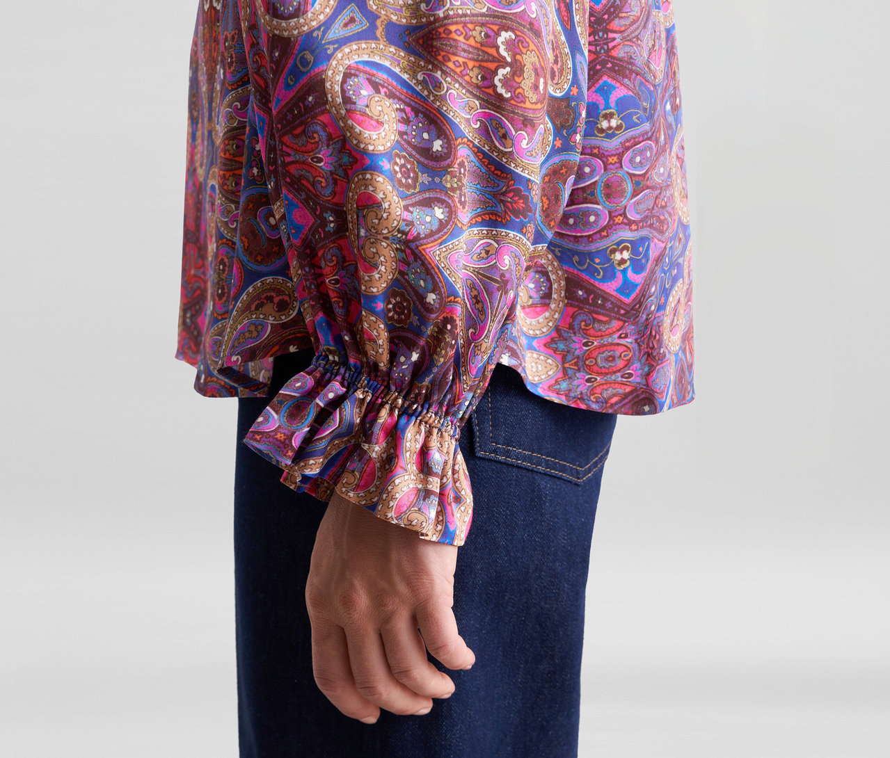 Person trägt eine bunte Bluse mit Paisley-Muster und Rüschenmanschette, dazu dunkle Jeans. Eine Hand ist sichtbar.