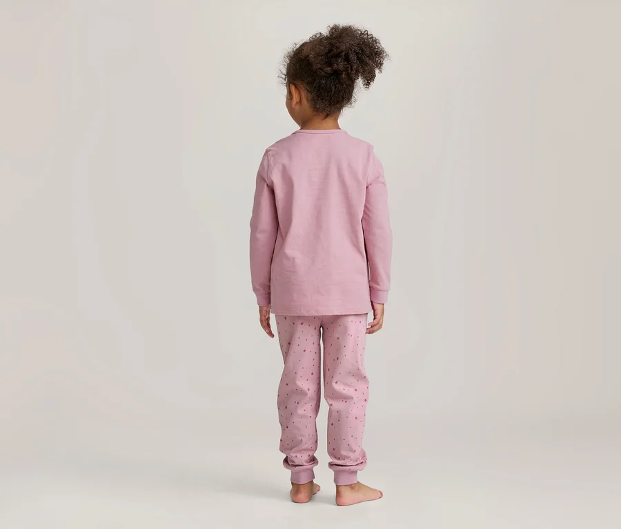 Junges Mädchen von hinten, in rosa langärmeligem Pyjama mit gemusterter Hose, barfuß vor hellem Hintergrund stehend.