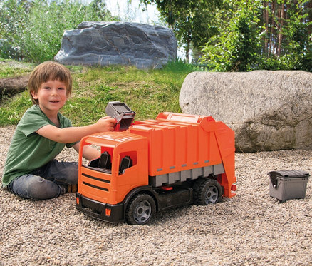 Ein lächelnder Junge sitzt auf Kies und spielt mit einem orangefarbenen GIGA TRUCKS Müllwagen, wobei er einen kleinen grauen Mülleimer in die Ladevorrichtung hält. Ein weiterer kleiner Mülleimer steht daneben.